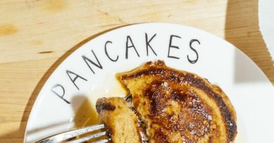 A fork cutting into a syrup-covered Lazy Weekend Pancakes stack on a plate that says “PANCAKES GO HERE!” rests on a wooden surface.