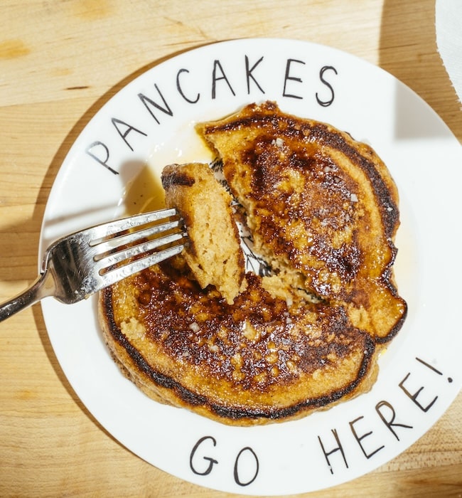 A fork cutting into a syrup-covered Lazy Weekend Pancakes stack on a plate that says “PANCAKES GO HERE!” rests on a wooden surface.