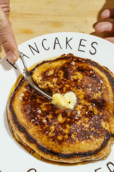 A hand uses a spoon to spread melting butter on a thick pancake with syrup, served on a plate labeled "PANCAKES"—the perfect treat for Lazy Weekend Pancakes.