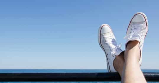 A person with crossed legs wearing white men's summer footwear rests their feet on a railing, overlooking a calm ocean under a clear blue sky.