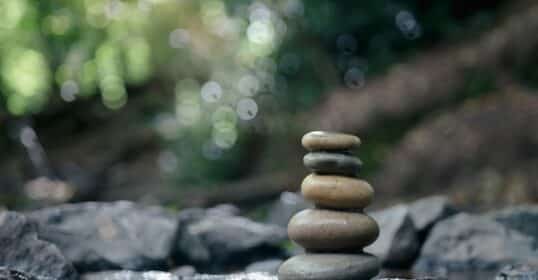 Five smooth stones stacked in a vertical pile on wet rocks beside a shallow stream, with blurred greenery in the background.
