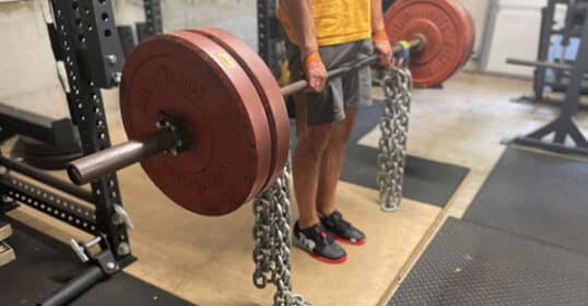 A person lifts a barbell loaded with red weight plates and heavy chains, using accommodating resistance in a gym setting.