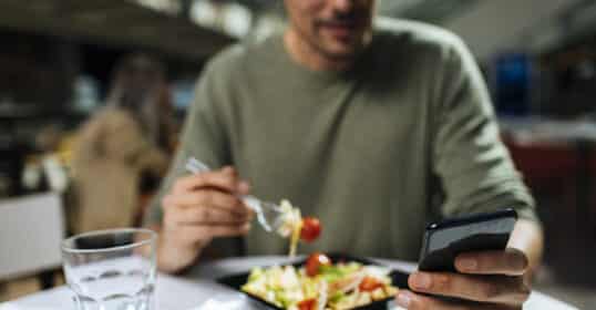 A person sits at a table eating a salad and using a smartphone, perhaps messaging their personal nutrition coach for tips, with a glass of water nearby. The focus is on their hands and the plate of food.