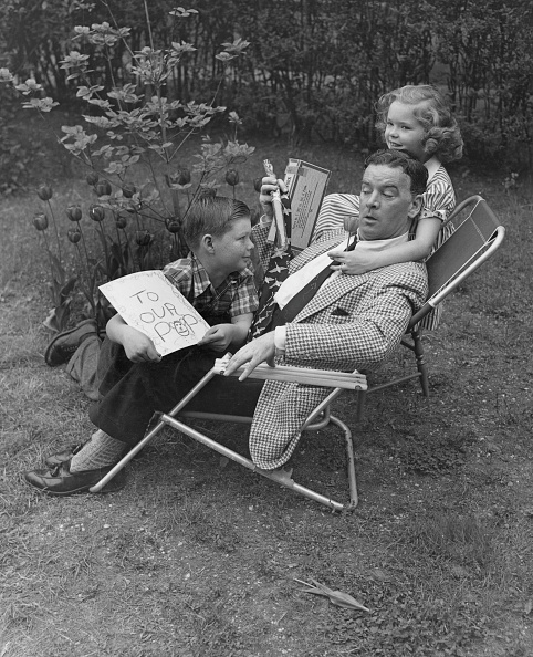 A man in a checkered suit sits in a tipped-over lawn chair while a boy shows him a card and a girl hangs on his shoulders, capturing the joy of Father’s Day in a lively garden setting.