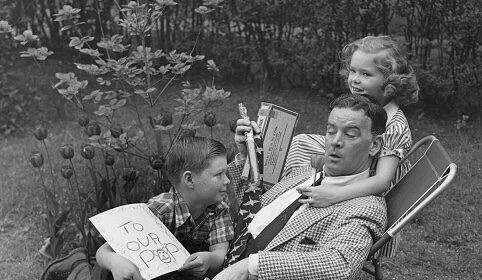A man sits in a garden chair holding a newspaper, while a boy shows him a card and a girl hugs him from behind, both children smiling—capturing the joy of Father’s Day and heartfelt Father’s Day gifts.