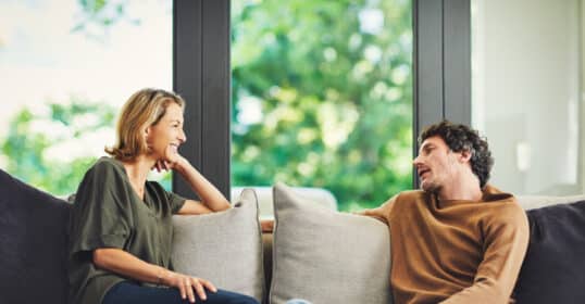 Two people sit on a sofa facing each other, engaged in conversation during a Marriage Checkup, with large windows and greenery visible in the background.