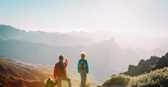 Three people with backpacks stand on a mountain trail, overlooking a scenic valley bathed in sunlight. One, guided by fatherly instinct, points out the view—launching kids into moments of wonder and discovery.