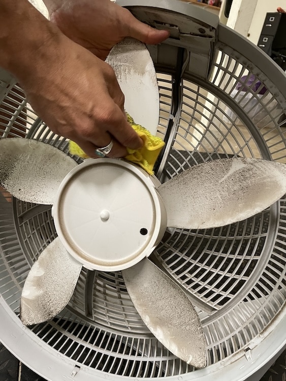 A person uses a yellow cloth to clean the dusty blades of a disassembled fan, showing an easy way to tackle Box Fan Cleaning.