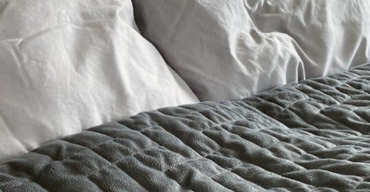 Close-up of a bed with two white pillows and a textured, dark gray weighted blanket spread across the mattress, highlighting the benefits of weighted blankets for restful sleep.