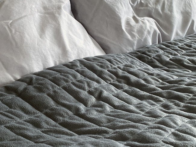 Close-up of a bed with two white pillows and a textured, dark gray weighted blanket spread across the mattress, highlighting the benefits of weighted blankets for restful sleep.