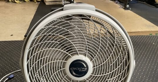 A Cyclone brand box fan with a circular grill sits on a rubber gym floor, plugged in and ready for use—perfect timing if you need tips on how to clean a box fan after an intense workout session.