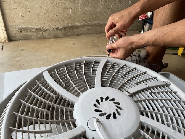 A person uses a screwdriver to remove screws from the front grill of a large box fan on the garage floor, showing how to clean a box fan effectively.