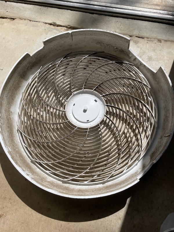 A dirty, white plastic fan cover with a circular metal grille sits on a concrete floor in sunlight—perfect for anyone learning how to clean a box fan.
