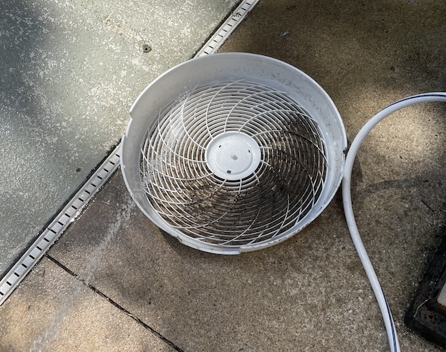 A white, round fan cover with visible dirt sits on a concrete surface next to a white cord and a textured glass or plastic panel, ready for box fan cleaning.
