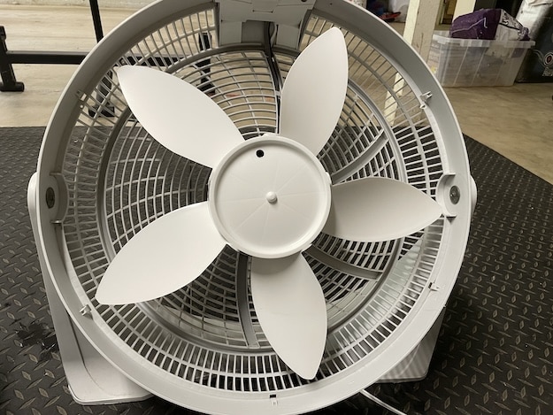 A close-up view of a large, white floor fan with five blades, positioned on a metal-patterned surface in an indoor setting—ideal for illustrating how to clean a box fan.