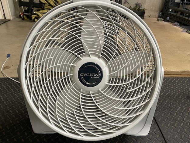 A white Cyclone brand box fan sits on a rubber mat in a room with gym equipment and plants in the background—a great spot to remember regular box fan cleaning for fresh, cool air during your workouts.