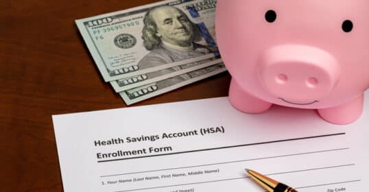 A Health Savings Account enrollment form sits on a desk next to a pen, three $100 bills, and a pink piggy bank—ready to help you build wealth for your six-figure future.