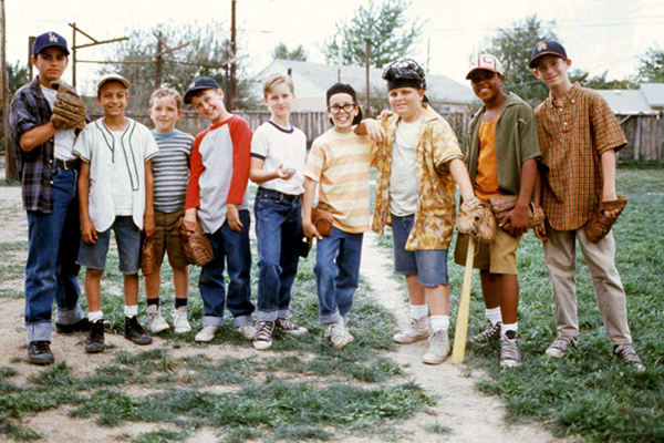 Ten boys stand in a row on a baseball field, some holding gloves and one holding a bat, posing for a group photo on a sunny day.