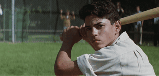 A boy with dark hair holds a baseball bat, preparing to swing, while standing on a grassy field near a chain-link fence.