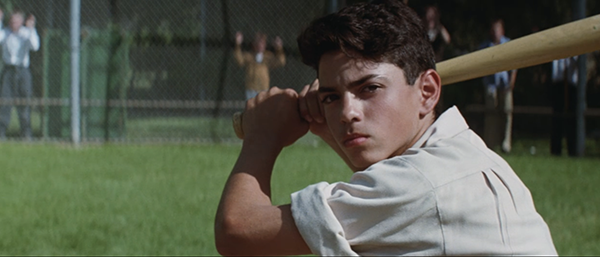 A boy with dark hair holds a baseball bat, preparing to swing, while standing on a grassy field near a chain-link fence.