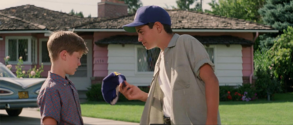 A boy in a blue cap hands another boy a baseball cap outside a suburban house with a vintage car parked nearby.