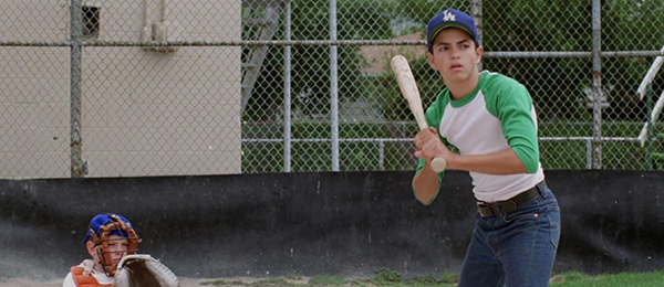 A teenage boy in a green and white shirt prepares to bat at a baseball field, while a catcher kneels behind him in full gear.