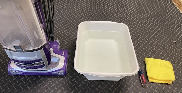 A vacuum cleaner, a white basin filled with water, a screwdriver, and a yellow cloth are placed on a patterned floor—essential tools when learning how to clean a box fan effectively.