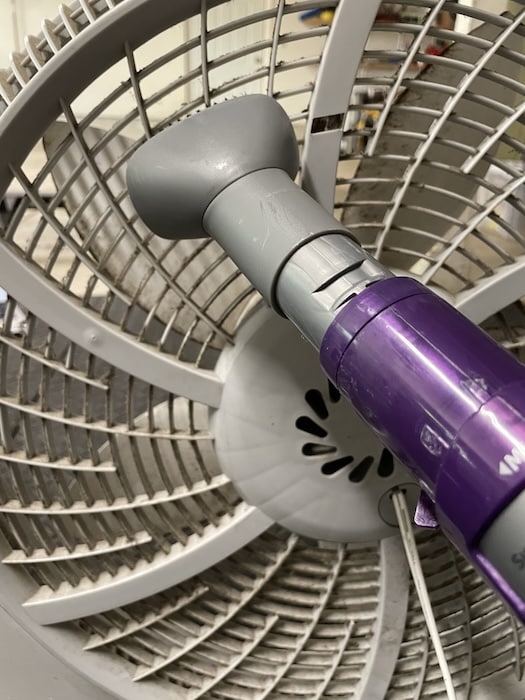 A handheld vacuum cleaner nozzle is shown demonstrating how to clean a box fan by removing dust from the grill of a large circular fan.