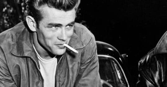 A young man with effortless style leans forward, wearing a white t-shirt and jacket, a cigarette in his mouth; a car is visible in the background.