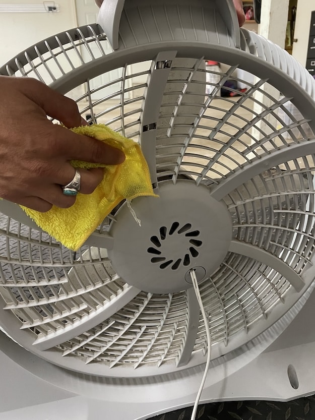 A person demonstrates how to clean a box fan by wiping the back grill of a white electric fan with a yellow cloth.