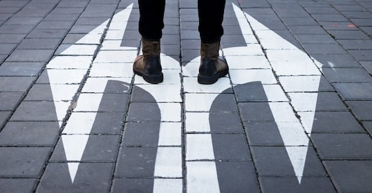 Person standing on gray pavement at a fork in a white painted directional arrow, symbolizing a big decision or choice ahead.