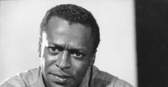 Black and white photo of a man with short hair, wearing an OCBD shirt buttoned up, looking directly at the camera with a serious expression.