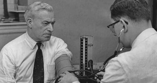 A doctor measures an older man's blood pressure using a manual sphygmomanometer and stethoscope, providing essential care for men's health in a clinical setting.