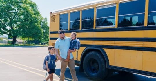 An adult dad walks with two children wearing backpacks toward a yellow school bus parked in a lot on a sunny, back-to-school day.