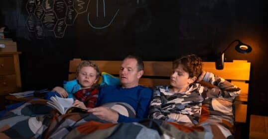 An adult and two kids sit in bed under a patterned duvet, reading aloud from classic chapter books, with a wall light and chalk drawings visible in the background.