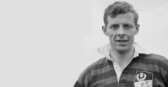 A man wearing a striped rugby shirt with an emblem stands outdoors, looking at the camera, against a plain light background—a classic look in men's fashion.