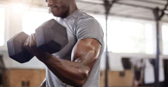 A person in a gray shirt lifts a dumbbell with one arm in a gym setting, showcasing strength and dedication to fitness.