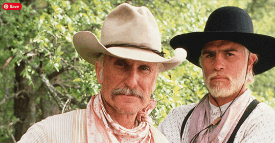 Two men in Western attire, resembling Gus and Woodrow, stand outdoors in front of leafy green trees, both wearing wide-brimmed hats and bandanas.