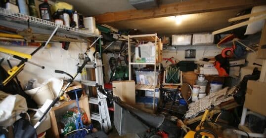 A cluttered garage with shelves full of boxes, tools, paint cans, and various stored items shows classic garage clutter. A lawnmower and a yellow toy vehicle are on the floor—perfect candidates for decluttering tips or things to get rid of.