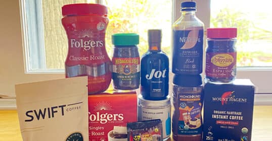 A variety of Instant Coffee products from different brands are displayed on a wooden table in front of a window.
