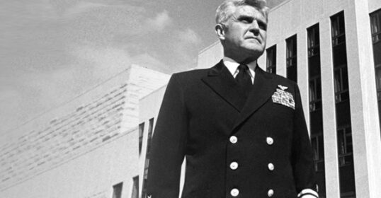 A man in a formal military uniform with medals stands outside a modern building, embodying the Stockdale Paradox as he looks into the distance, reflecting on tough times with unwavering resolve.