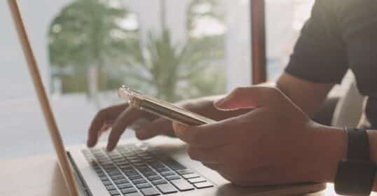 A person demonstrates task switching by using a smartphone in one hand while typing on a laptop with the other, seated at a desk near a window.