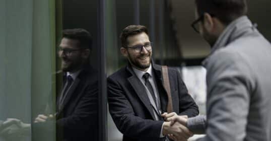 Two men in business attire shake hands and smile while standing outside near a glass building, showcasing how positive habits can help improve relationships—an approach inspired by Dale Carnegie’s timeless principles.