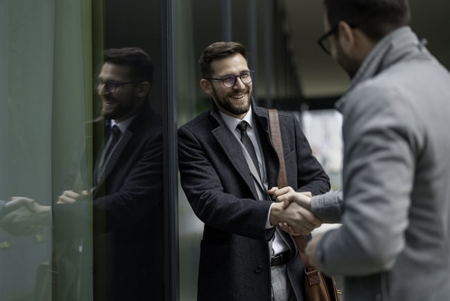 Two men in business attire shake hands and smile while standing outside near a glass building, showcasing how positive habits can help improve relationships—an approach inspired by Dale Carnegie’s timeless principles.