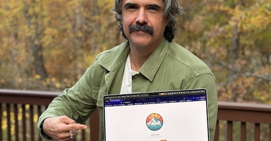 A person with a mustache points to a laptop screen displaying the Art of Manliness website and colorful mountain logo, standing outdoors among trees—inviting viewers to support their journey or join the free newsletter.