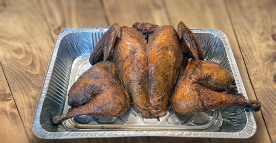 A whole smoked turkey, spatchcocked and browned using our smoked turkey recipe, sits in a disposable aluminum roasting pan on a wooden surface.