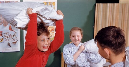 Three children in pajamas have a pillow fight in a room, with posters on the wall behind them, as their favorite podcast plays softly in the background.