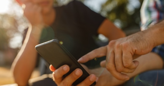 Two people outdoors looking at a smartphone, with one person pointing at the screen and the other resting their chin on their hand—perhaps discussing parenting rules for raising kids in today’s high-tech world.
